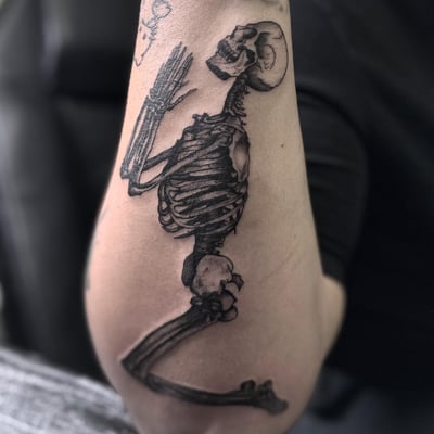 A black and grey skeleton tattoo in a kneeling position on the arm — by Maddie Ramirez, los angeles