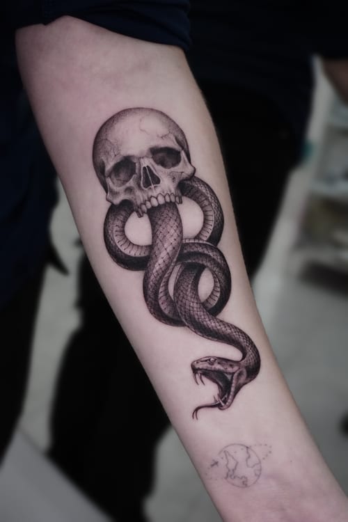 A realistic black and grey tattoo of a skull with a snake coiled through it on the forearm, featuring detailed shading and texture — by Jian Liang, los angeles