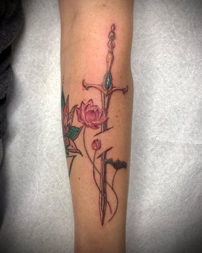 A delicate fine line tattoo of a sword intertwined with a lotus flower on the forearm, featuring colorful details — by Sandra Price, los angeles