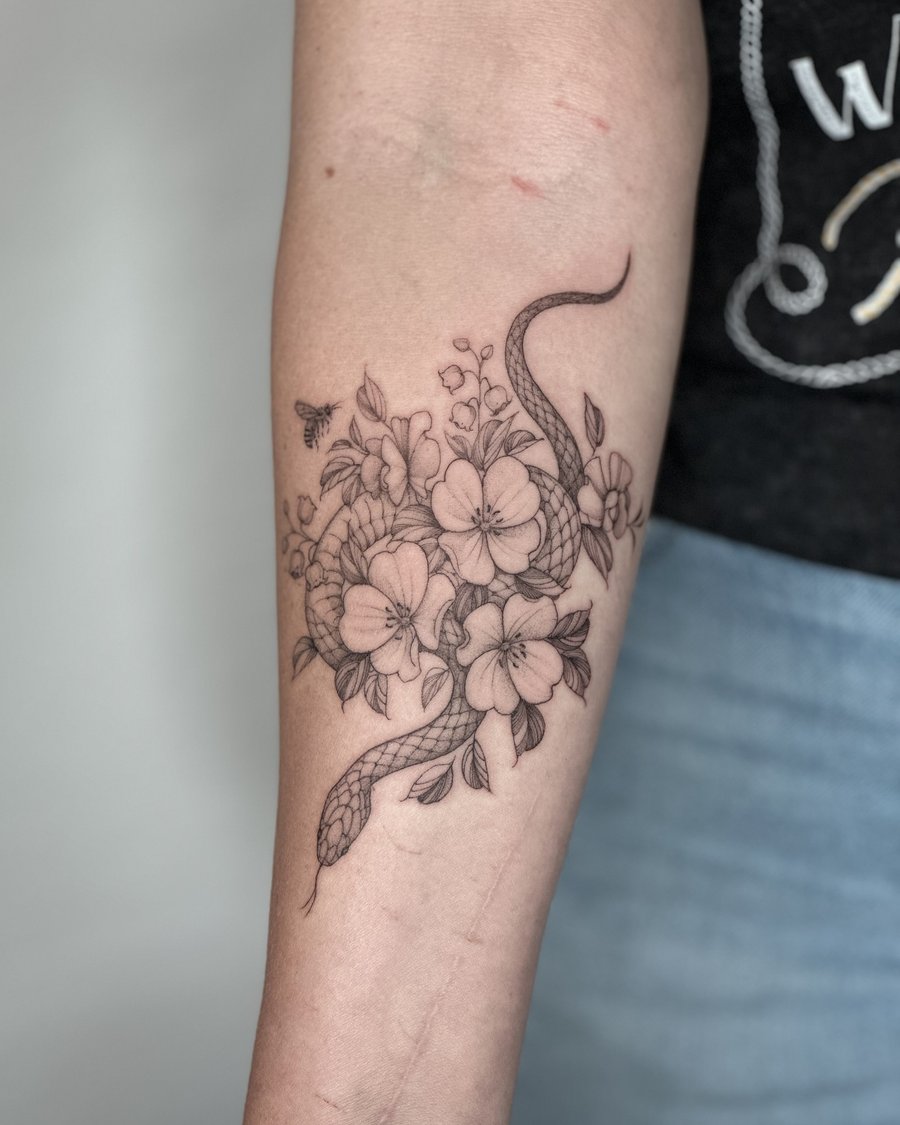 A delicate black and grey fine line tattoo of a snake intertwined with flowers on the forearm — by Melanie Benoit, los angeles