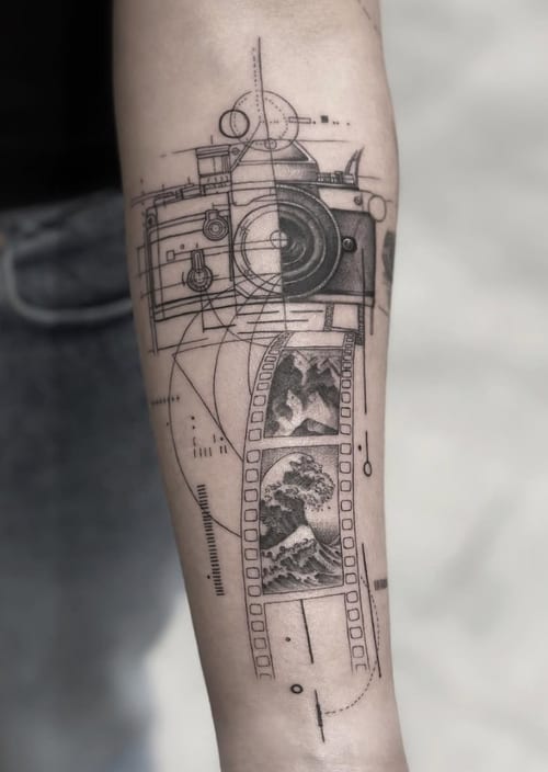 A black and grey geometric and illustrative tattoo on the forearm depicting a camera with film strips showing landscape scenes, featuring precise linework and detailed imagery — by Jian Liang, los …