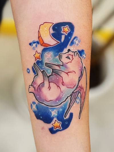 A colorful watercolor tattoo of a winged animal floating among stars and a crescent moon on the forearm, featuring vibrant blues, pinks, and oranges — by Tweetwee, los angeles
