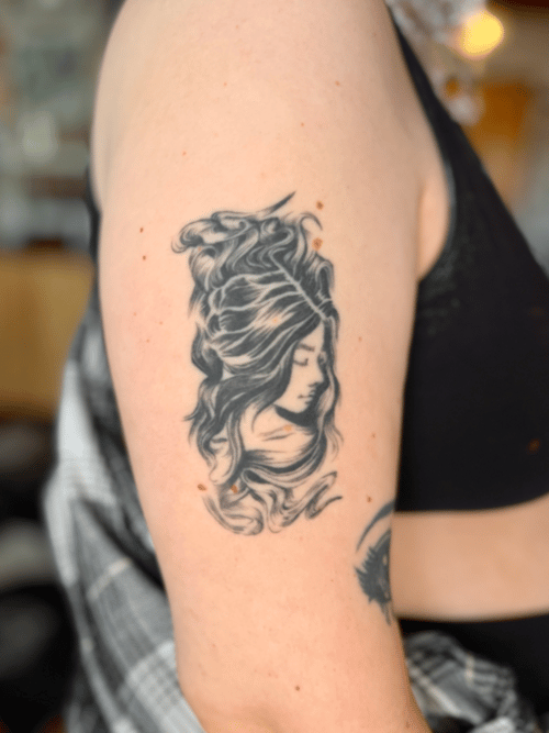 A black and grey realism tattoo of a woman's face with flowing hair on the upper arm, featuring soft shading and intricate details — by Dylan Aiello, los angeles