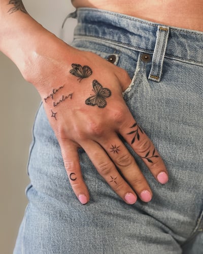 A delicate black and grey tattoo on the hand featuring fine line butterflies and script lettering, with minimalist symbols on the fingers — by Melanie Benoit, los angeles
