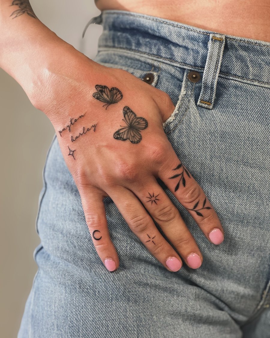 A delicate black and grey tattoo on the hand featuring fine line butterflies and script lettering, with minimalist symbols on the fingers — by Melanie Benoit, los angeles