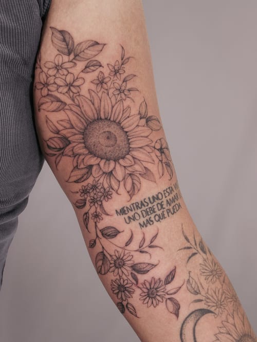 A delicate black and grey fine line tattoo of a sunflower and floral arrangement with script on the arm — by Melanie Benoit, los angeles