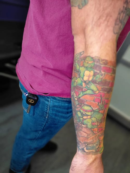 A colorful illustrative tattoo on the forearm depicting cartoon-like animals with a brick wall background — by anoetic.designs.ultd, los angeles