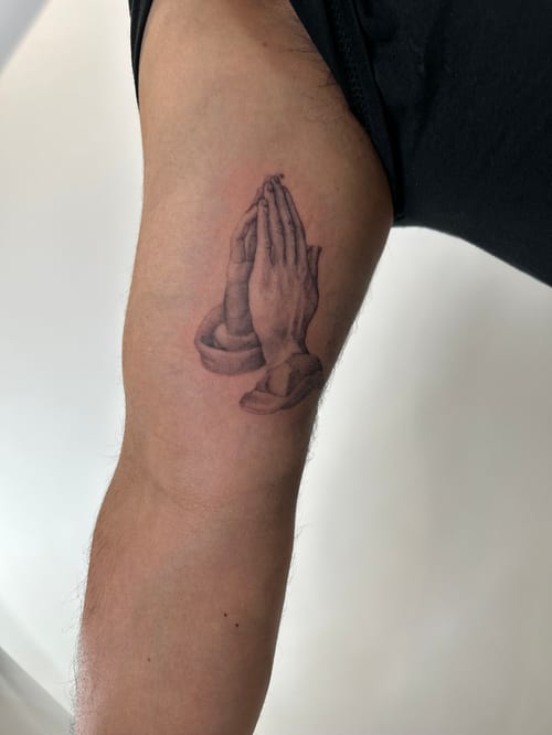 A realistic black and grey tattoo of praying hands on the upper arm, symbolizing faith and spirituality — by Brian Kies, los angeles
