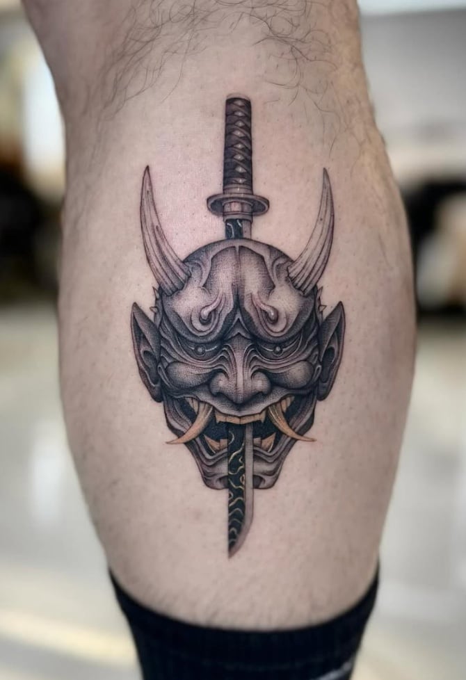 A black and grey Japanese style tattoo of a demon mask with a sword running through it, located on the calf with intricate shading and detail — by otaybeatdown, los angeles