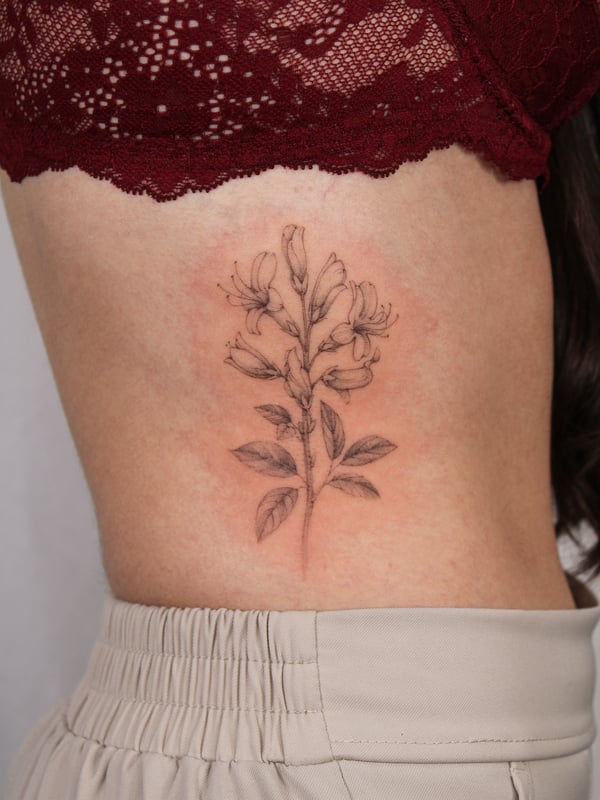 A delicate black and grey fine line flower tattoo on the rib, showcasing a botanical style — by Melanie Benoit, los angeles