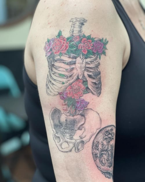 A mixed color and black and grey tattoo of a ribcage with colorful flowers on the upper arm, combining skeletal and floral elements — by Sandra Price, los angeles