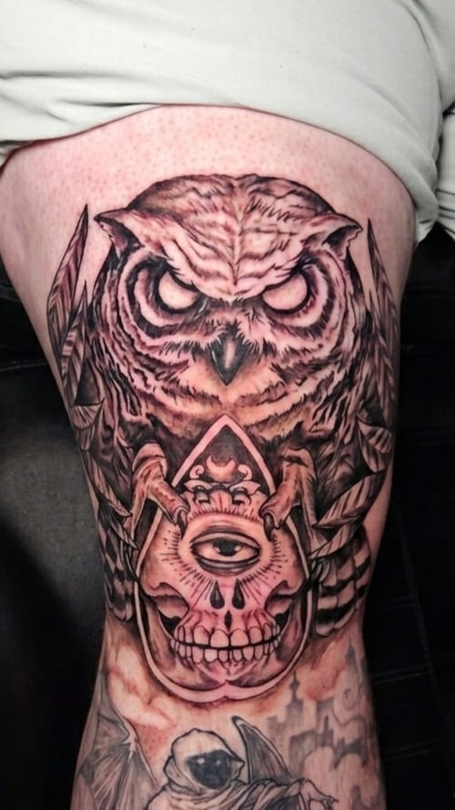 A black and grey tattoo on the thigh featuring an owl perched above a skull with an eye in the center, rendered in a neo-traditional style with dark art elements — by Dretty, los angeles