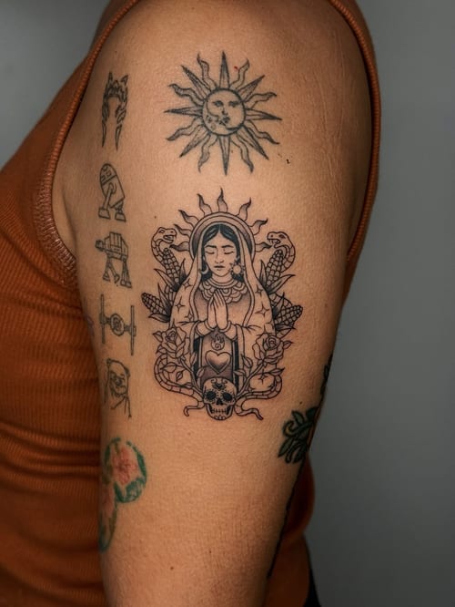 A black and grey tattoo on the upper arm featuring a praying woman surrounded by snakes, a skull, and roses, with a sun above — by lunayflorestattoo, los angeles