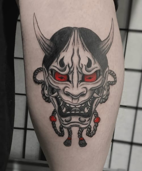 A Japanese-style demon mask tattoo on the calf with bold black outlines and red accents in the eyes and beads — by Shelby Bianco, los angeles