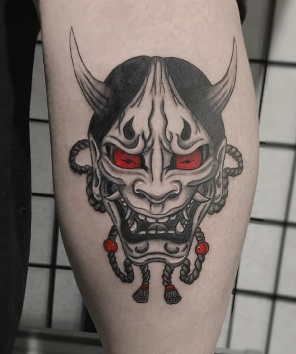 A Japanese-style demon mask tattoo on the calf with bold black outlines and red accents in the eyes and beads — by Shelby Bianco, ventura, ca