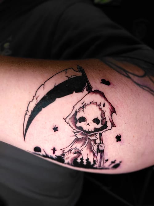 A black and grey neo-traditional tattoo of a cartoonish grim reaper with a scythe on the upper arm, featuring bold lines and dark shading — by anoetic.designs.ultd, los angeles