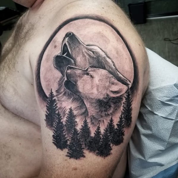 A black and grey realism tattoo of two wolves howling with a forest background on the upper arm, featuring detailed shading and texture — by Dretty, long beach, ca