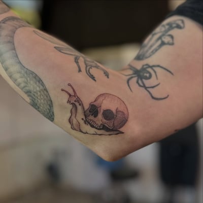 A dark blackwork tattoo of a snail with a skull on the inner arm — by Maddie Ramirez, los angeles