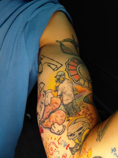 A colorful illustrative tattoo on the upper arm featuring a heart, skulls, and various script elements in a pop art style — by anoetic.designs.ultd, los angeles