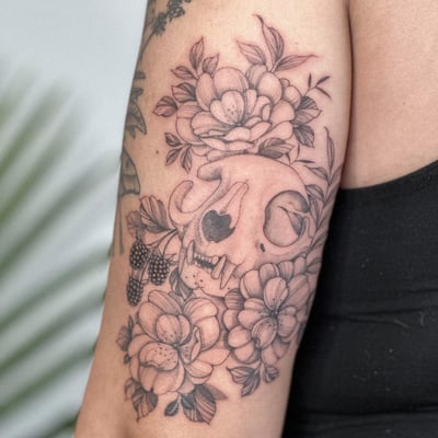A delicate black and grey tattoo of an animal skull surrounded by flowers on the upper arm — by Melanie Benoit, los angeles