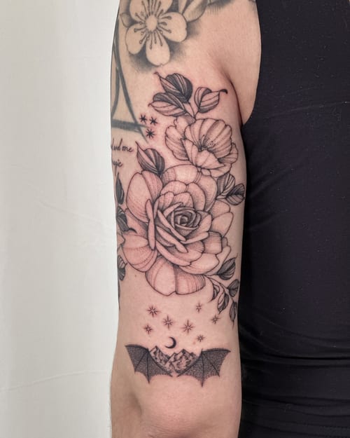 A delicate black and grey tattoo featuring a rose with leaves and a bat with a moon and stars on the upper arm — by Melanie Benoit, los angeles