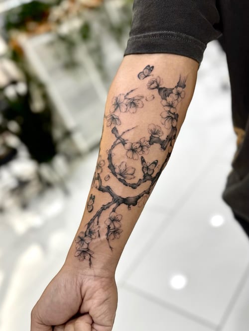 A black and grey fine line tattoo of cherry blossoms and butterflies on the forearm, featuring delicate linework and shading — by otaybeatdown, los angeles