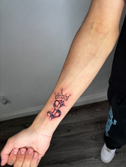 A color illustrative tattoo of a stylized letter 'S' with a crown on the forearm, featuring bold outlines and shading — by Edgar Armas Rodriguez, los angeles