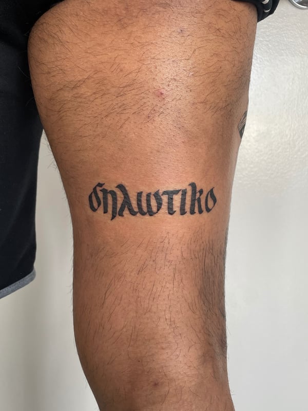 A bold black script lettering tattoo on the thigh in a gothic style — by Brian Kies, los angeles