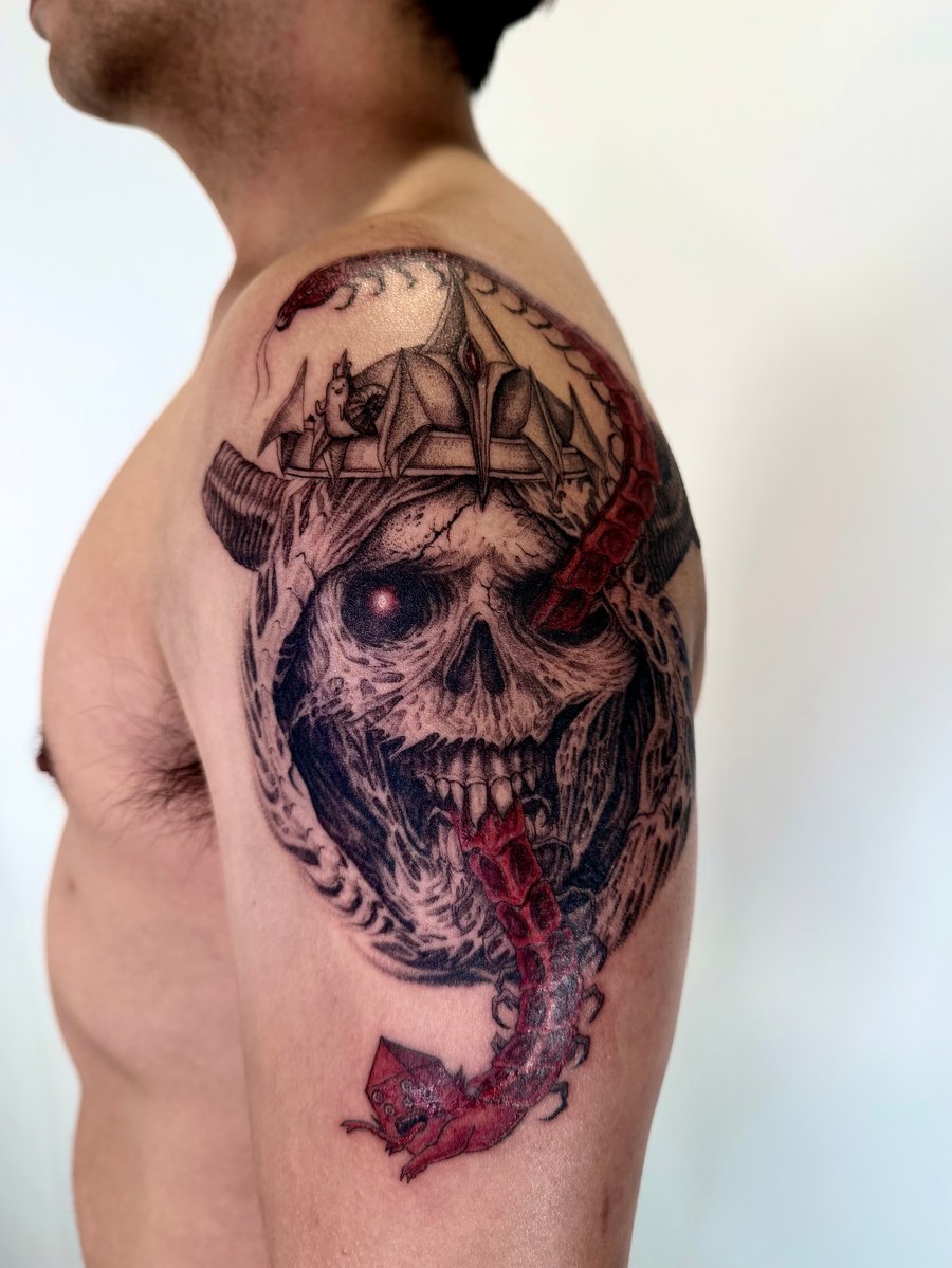 A dark and illustrative tattoo on the upper arm featuring a skull with a crown and a red snake. The design is primarily black and grey with striking red accents — by zitronxus, los angeles