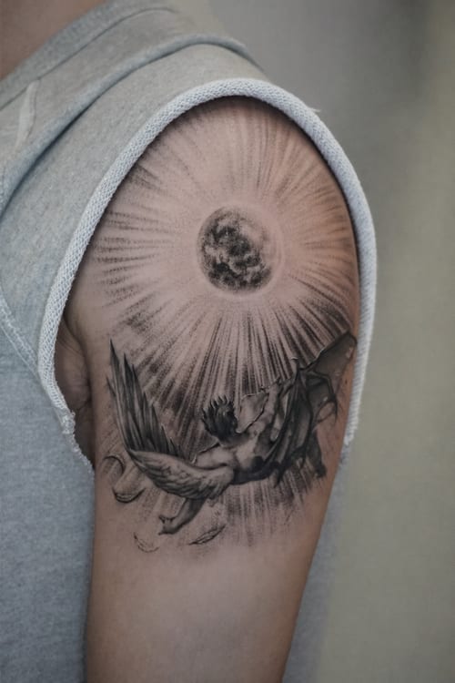 A black and grey tattoo on the upper arm depicting an angel reaching towards a detailed moon with radiant beams — by Jian Liang, los angeles