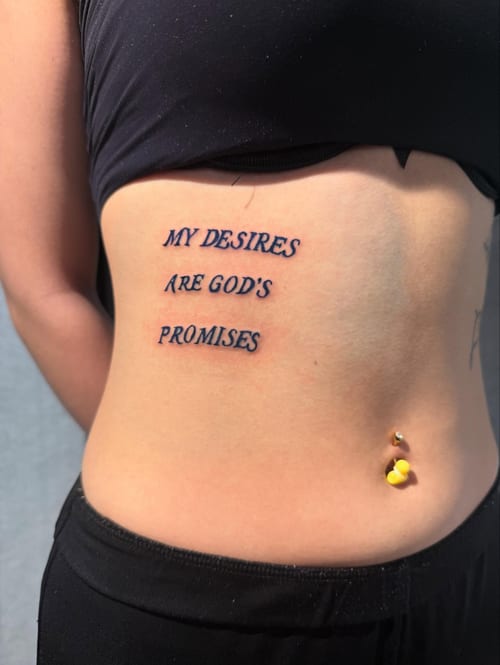 A black script lettering tattoo on the rib reading 'My desires are God's promises' in a minimal style — by Shelby Bianco, los angeles