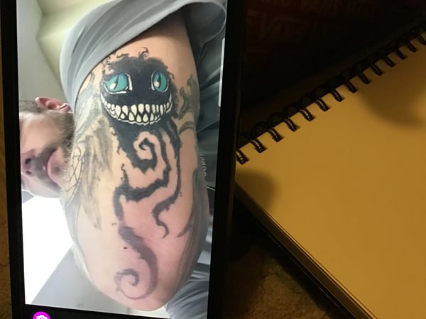 A mixed color illustrative tattoo of a grinning face with swirling patterns on the upper arm, featuring prominent black ink with striking blue eyes — by anoetic.designs.ultd, redlands, ca