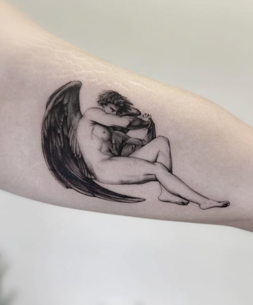 A black and grey realism tattoo of an angel sitting with wings folded, located on the inner arm with detailed shading — by Jian Liang, los angeles