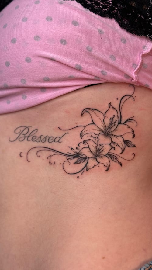 A black and grey fine line tattoo on the rib featuring the word 'Blessed' with floral elements, showcasing delicate script and flower details — by NJ TATTOOZ, los angeles