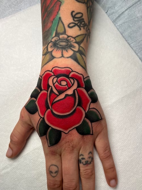 A vibrant American Traditional style rose tattoo on the hand, featuring bold outlines and classic red and green colors — by Megan Rose, los angeles