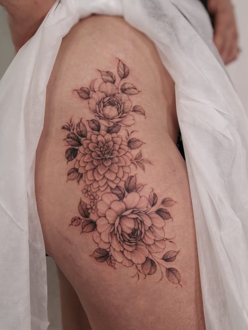 A delicate black and grey floral tattoo on the thigh featuring detailed flowers and leaves — by Melanie Benoit, los angeles