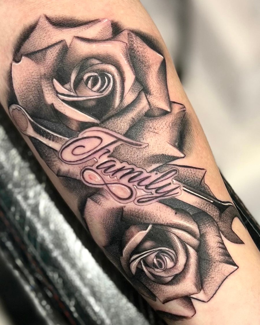 A black and grey tattoo on the forearm featuring two realistic roses with the word 'Family' in elegant script across them — by Josue Bravo, los angeles