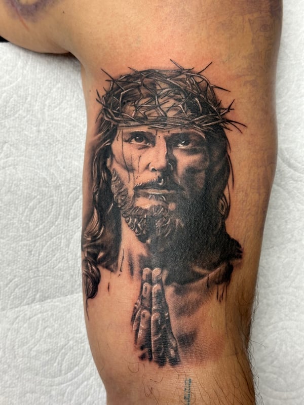 A black and grey realism portrait of Jesus Christ with a crown of thorns on the upper arm, featuring detailed shading and lifelike depiction — by Humilde tattoos, los angeles