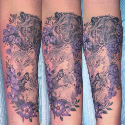 A realistic and colorful tattoo of wolves surrounded by purple flowers on the forearm — by Sandra Price, los angeles