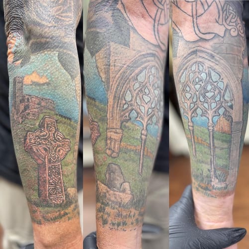 A colorful tattoo of a Celtic cross and ruins on the forearm, featuring realistic architectural details and a landscape background — by Sandra Price, los angeles
