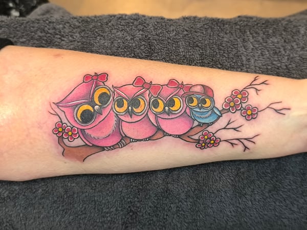 A whimsical and colorful tattoo of four cartoon owls sitting on a branch with flowers, located on the forearm — by Samuel Connelly, san diego, ca