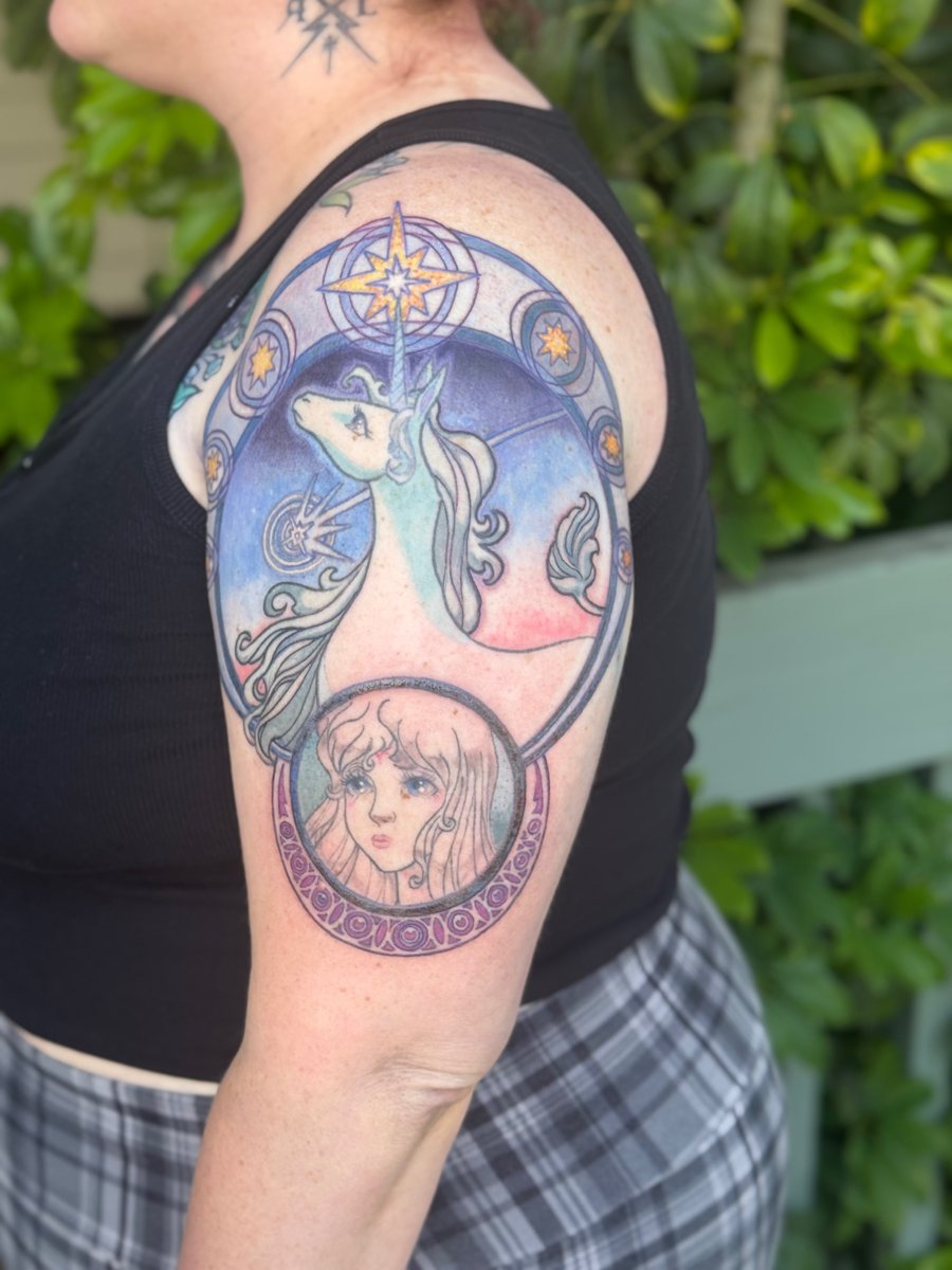 A colorful unicorn and woman's face tattoo on the upper arm with celestial elements in a whimsical and delicate style — by Sandra Price, los angeles