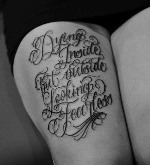 A bold black and grey script lettering tattoo on the thigh featuring a quote with intricate swirls — by Johnny Ayala, los angeles
