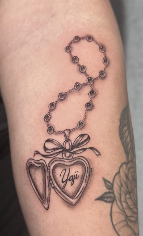 A black and grey fine line tattoo of a heart-shaped locket with a chain, featuring script inside, on the arm — by Shelby Bianco, los angeles