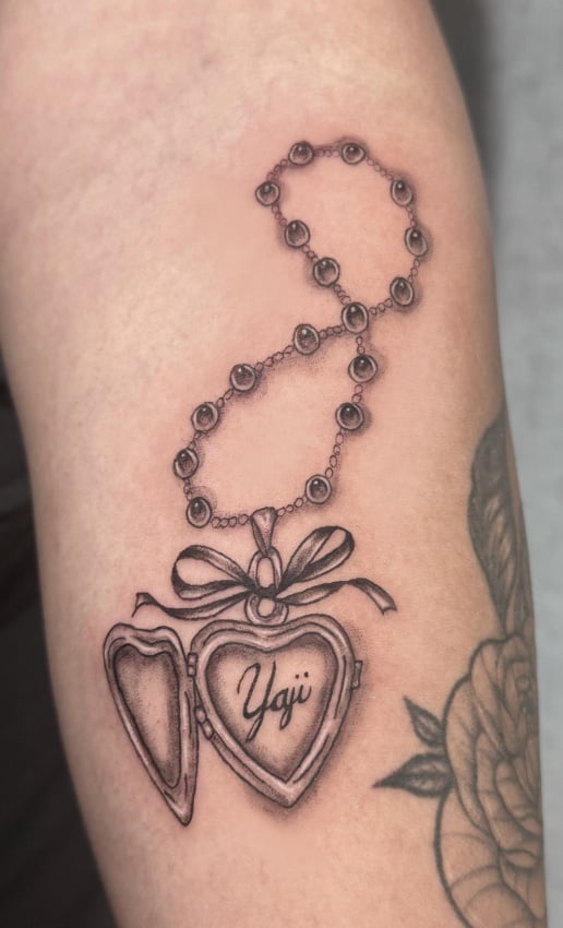 A black and grey fine line tattoo of a heart-shaped locket with a chain, featuring script inside, on the arm — by Shelby Bianco, los angeles