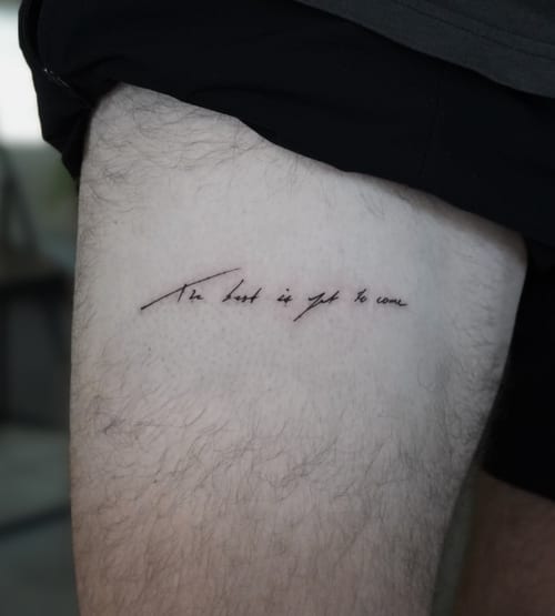 A minimalist black script tattoo on the thigh with the quote 'The best is yet to come' in elegant handwriting — by Jian Liang, los angeles