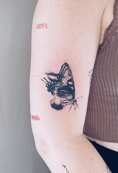 A black and grey tattoo on the upper arm depicting a realistic butterfly merging with a human face, showcasing intricate detail and shading — by Level tattoo, los angeles