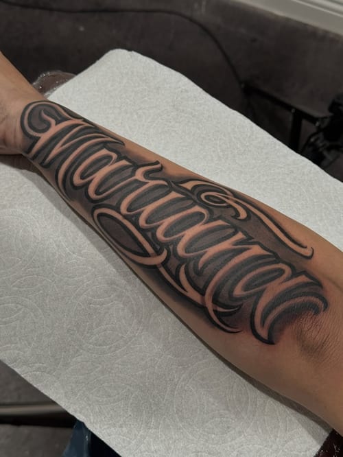A bold black and grey script lettering tattoo on the forearm — by Johnny Ayala, los angeles