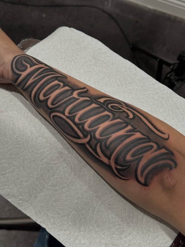 A bold black and grey script lettering tattoo on the forearm — by Johnny Ayala, fontana, ca