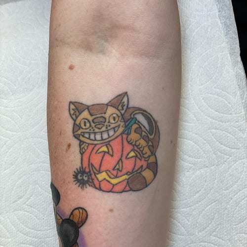 A whimsical and colorful tattoo of a cat sitting on a carved pumpkin, located on the forearm — by Sandra Price, los angeles